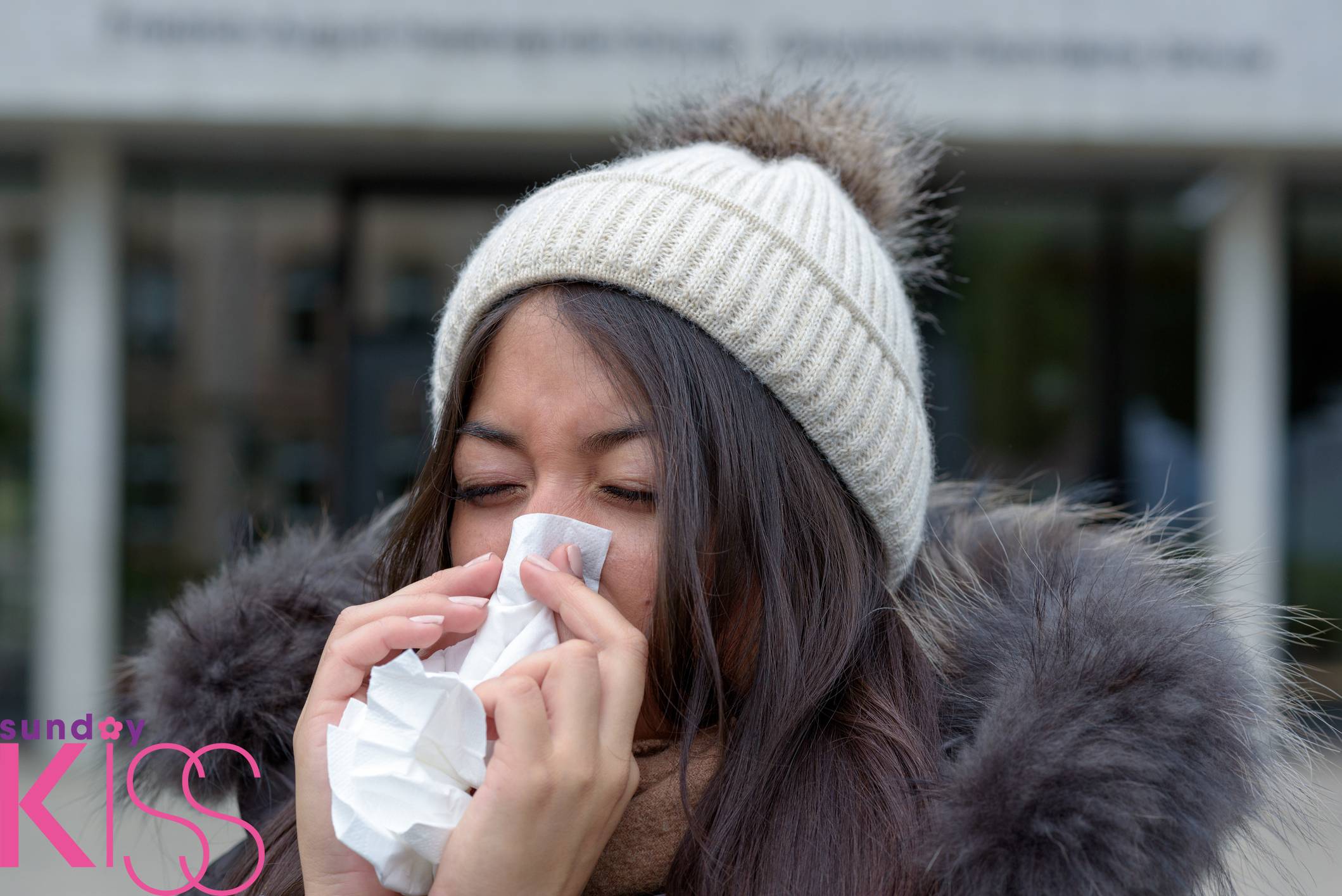 立冬湯水 Young woman with a seasonal winter cold or flu wearing a furry jacket and knitted cap blowing her nose on a white handkerchief outdoors on an urban street